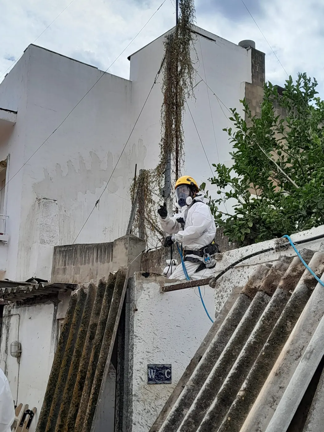 Operario con casco, buzo de protección y arnés anclado trabajando en altura sobre el muro de la cubierta de uralita
